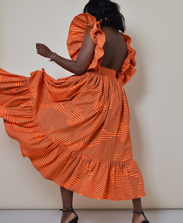 Model wearing The Kemist Barletta Backless Dress in Orange on a plain cream backdrop. 